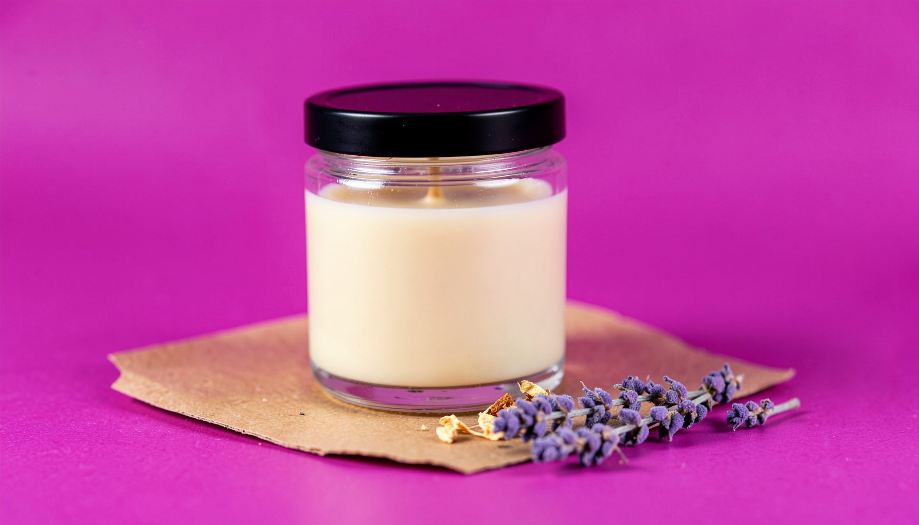 A softly lit close-up of a hand-poured soy candle in a clear glass jar with a black lid, resting on brown kraft paper beside a few dried flower petals and a sprig of lavender, warm cream and blush tones.