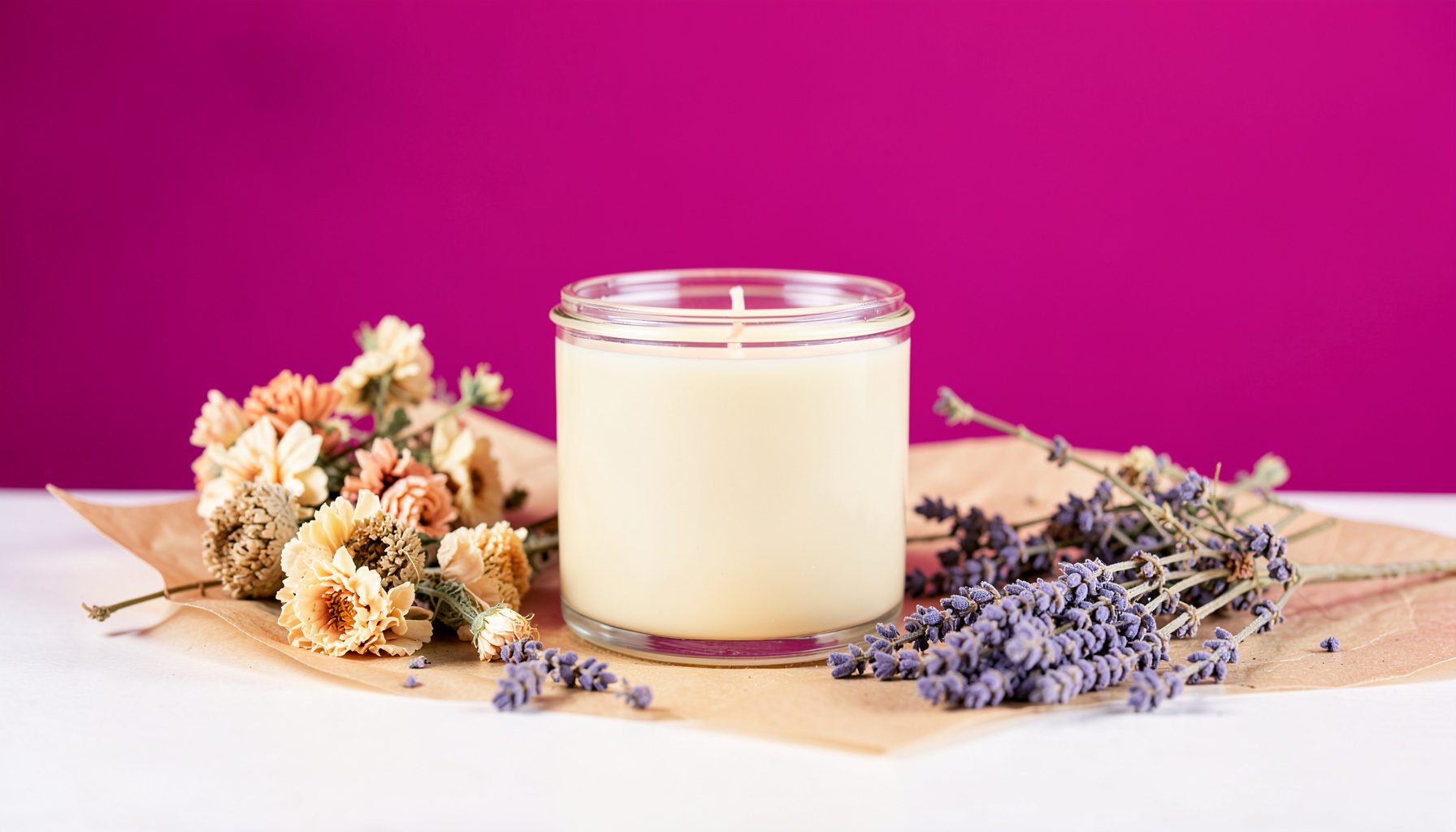 A softly lit studio table with a hand-poured soy candle in a clear jar, surrounded by dried flowers and sprigs of lavender on brown paper. Warm cream and blush tones.