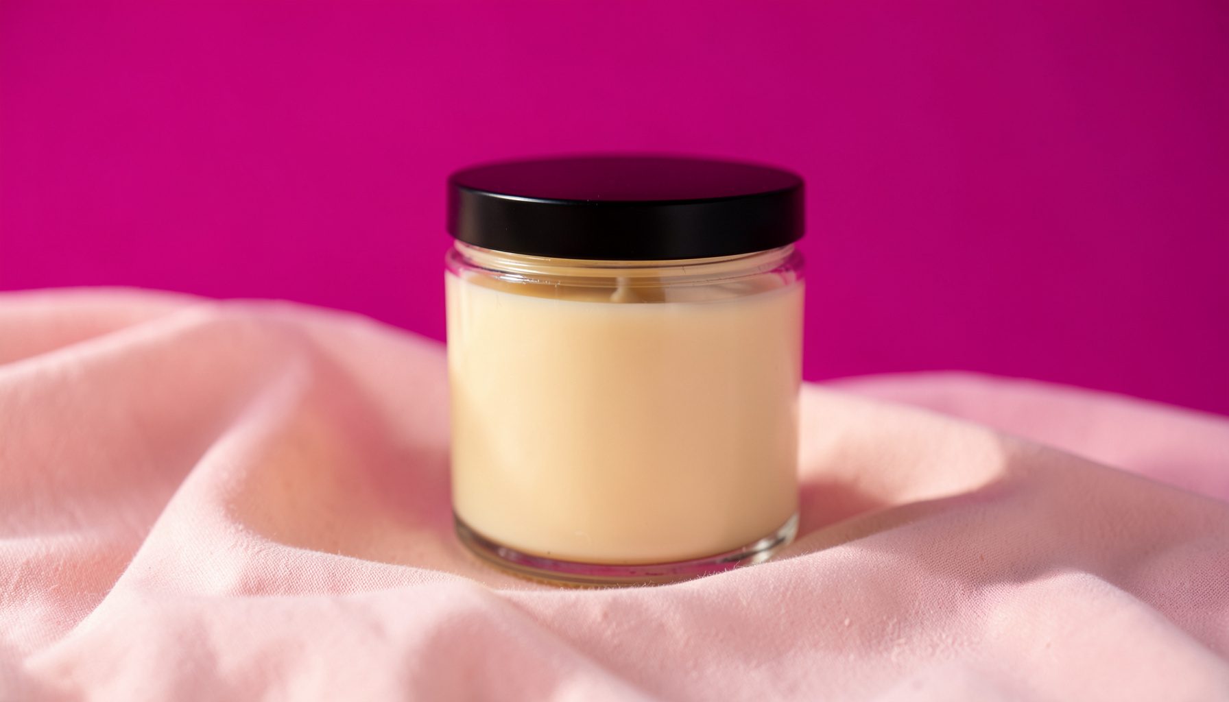 A single hand-poured soy candle in a clear glass jar with a black lid, resting on a soft blush-pink fabric backdrop. Warm, diffused daylight wraps the scene in a gentle, romantic glow.