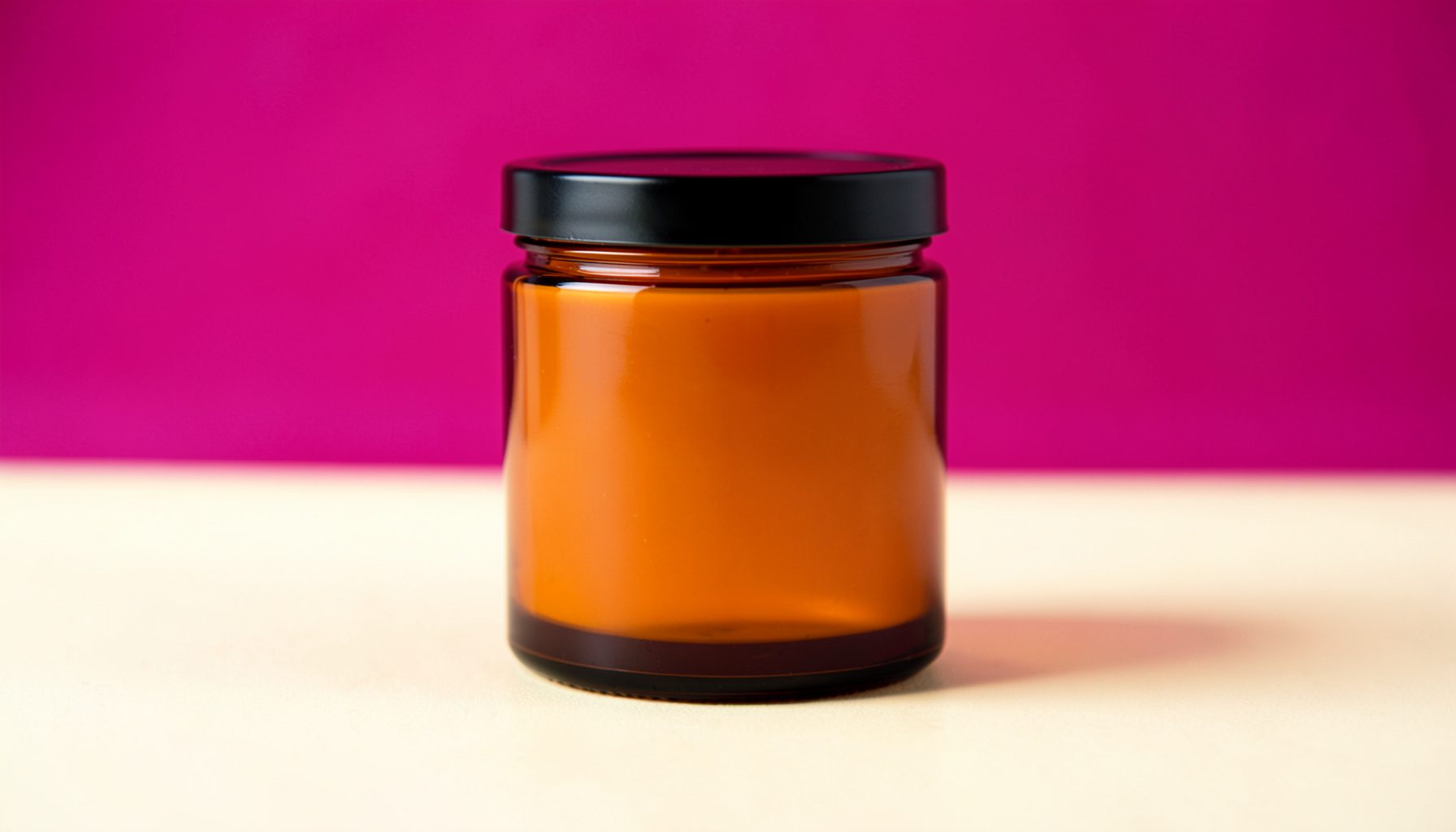 An amber glass jar candle with a black lid on a soft cream backdrop, warm rose and sandalwood tones in the surrounding atmosphere